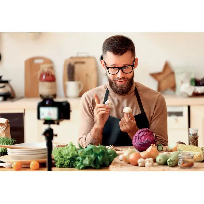 Bărbat cu barbă și ochelari ținând ciuperci albe în mână lângă legume proaspete și sort din bumbac organic 200 gr/m² beige