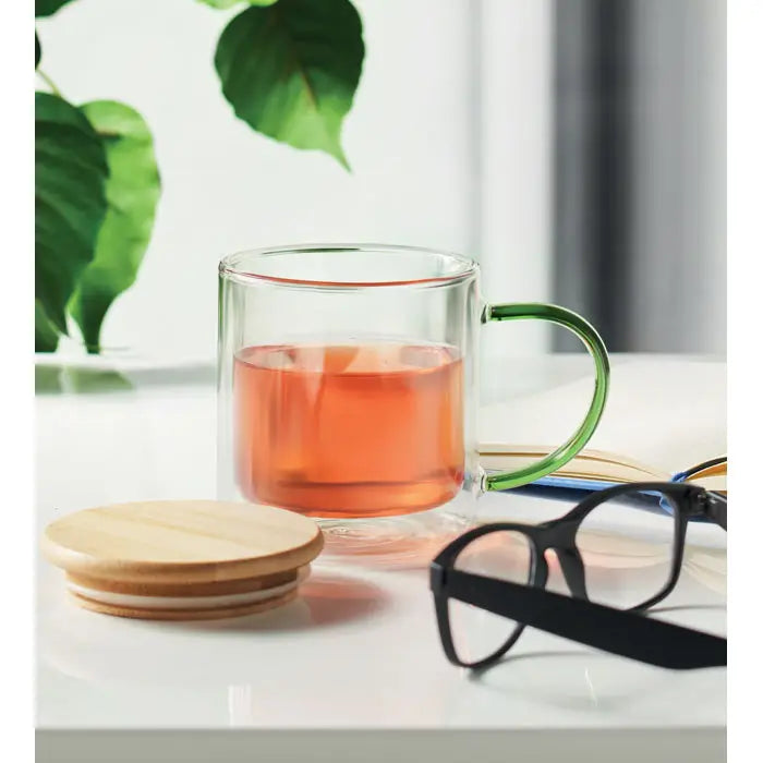 Cană din borosilicat cu perete dublu, mâner verde vibrant, umplută cu lichid de culoare chihlimbar, transparentă