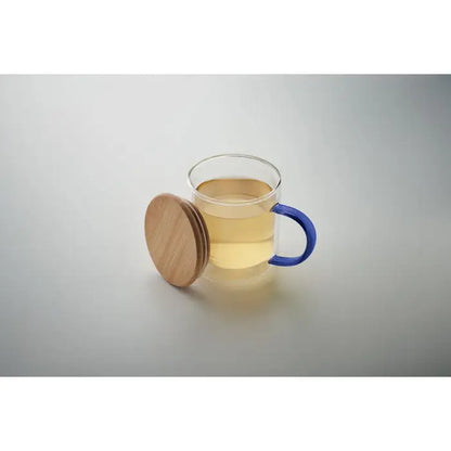 Cană din borosilicat cu perete dublu, mâner albastru și capac din lemn natural, umplută cu ceai auriu