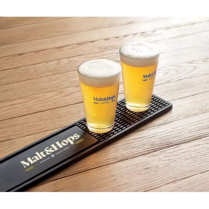 Două pahare de bere clare din sticlă cu spumă albă și logo malt&hops pe covoraș de bar din pvc galben