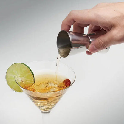 Jigger din otel inoxidabil argintiu folosit pentru a turna lichid clar in pahar martini garnisit cu lime si cireasa