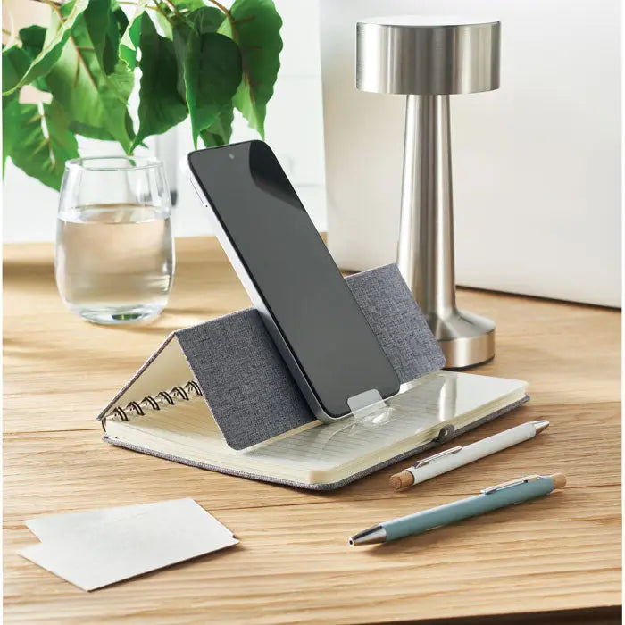 Notebook a5 cu suport pentru telefon din material textil gri ținând un smartphone negru