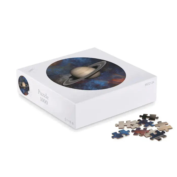 Cutie albă cu puzzle de 1000 de piese reprezentând Saturn cu inelele sale pe fundal cosmic și piese albastre și bej răspândite