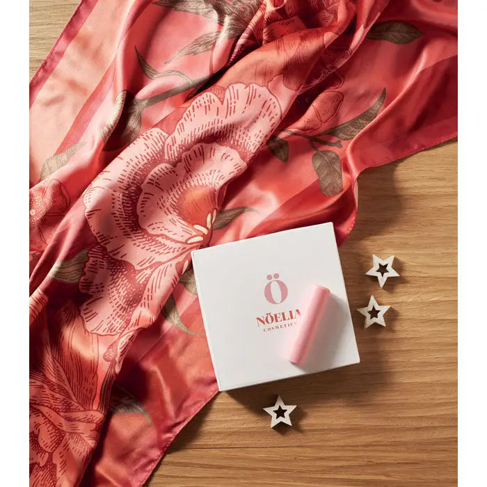 Un ruj roz deschis din poliester satinat reciclat lângă cutia albă minimalistă a brandului nöelia cosmetics