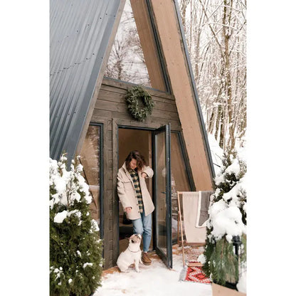 O femeie in palton bej sta la intrarea unei cabane in forma de a-frame in natura inzapezita lângă scaun de plaja din lemn