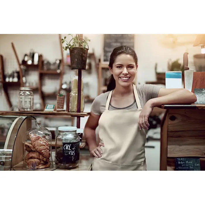 Sort din bumbac organic 200 gr/m² beige apron purtat de o femeie zâmbitoare în spatele unei tejghele de brutărie