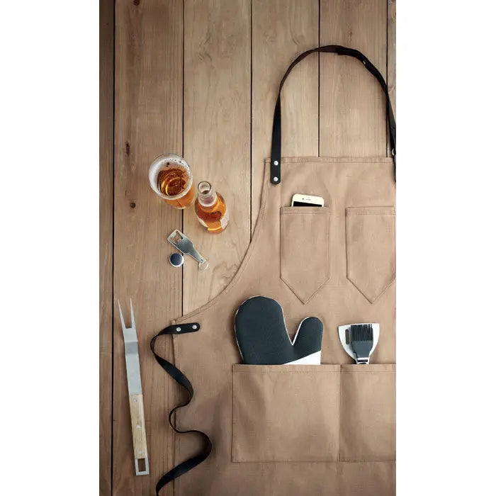Sort din piele tan canvas apron with black leather straps and multiple pockets holding barbecue tools